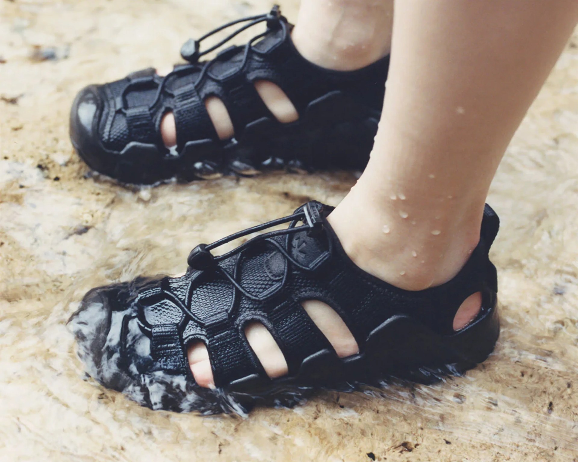 Black water shoes with mesh and adjustable laces worn in shallow flowing water.