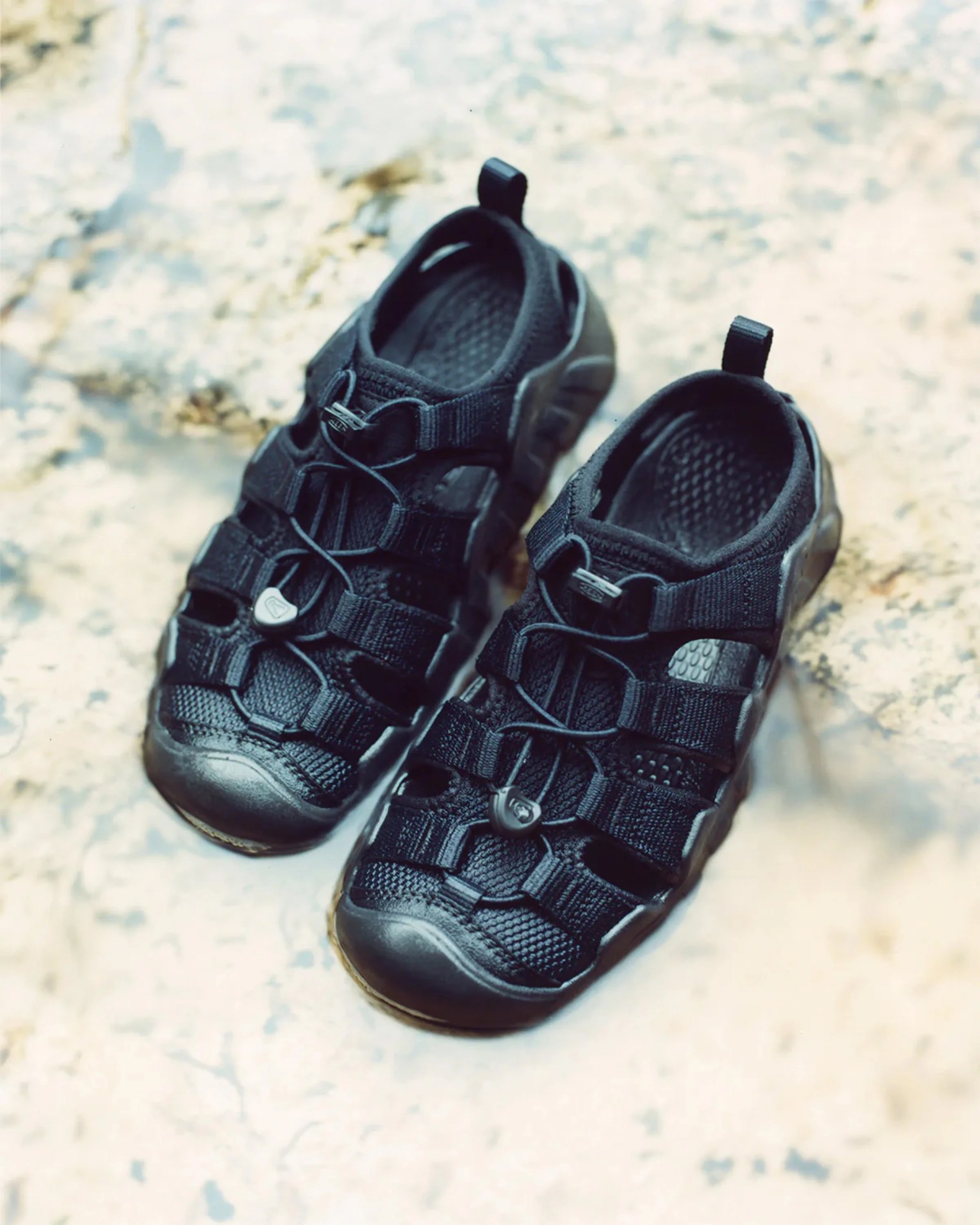 Pair of black outdoor water shoes with mesh and adjustable bungee laces on a stone surface.