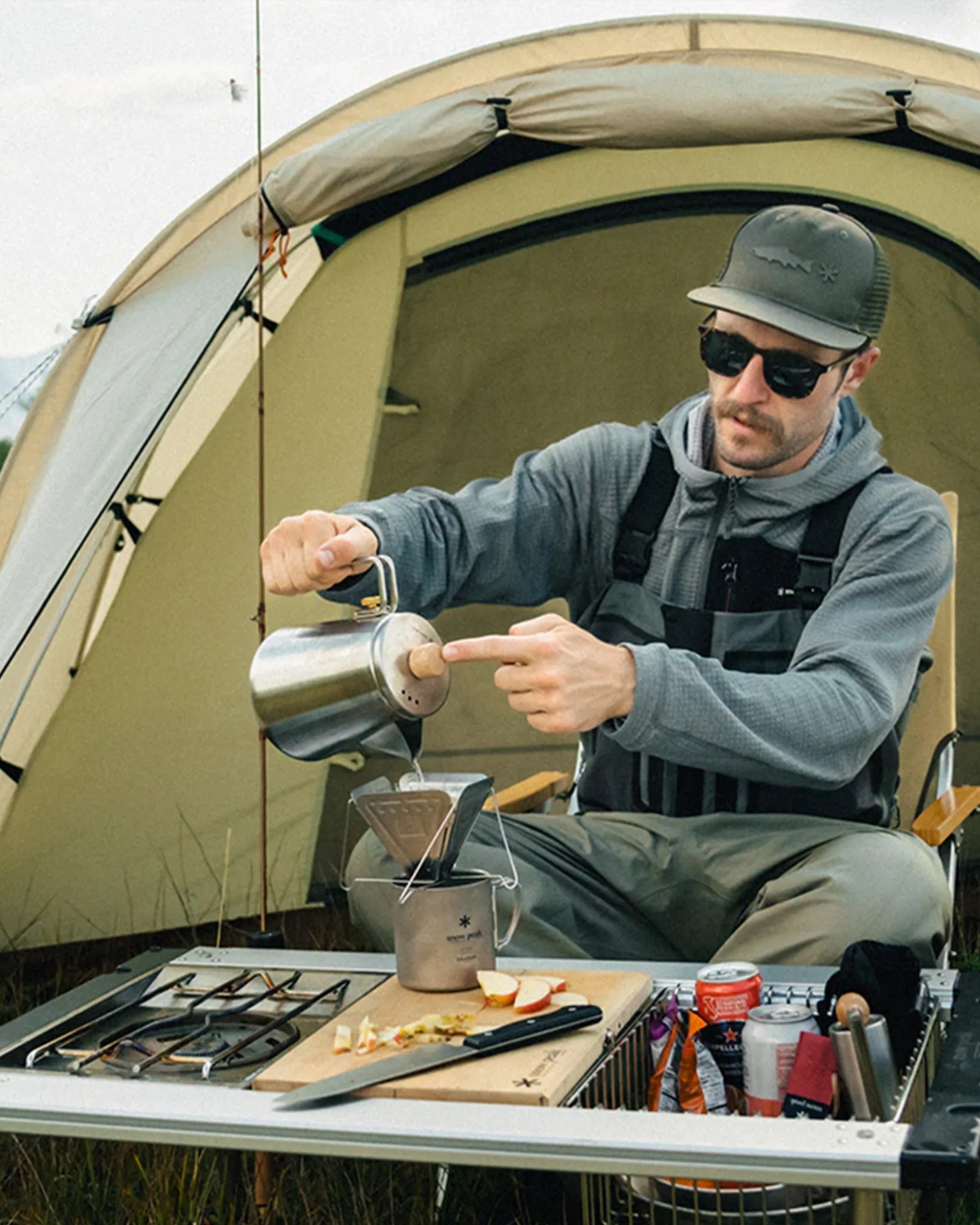Man wearing sunglasses and a cap pouring water from a metal kettle into a camping coffee dripper on a portable stove table.