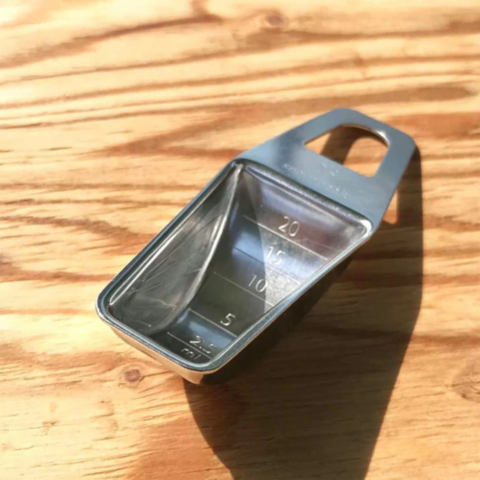 Small stainless steel measuring scoop with volume markings up to 20 ml on a wooden surface.