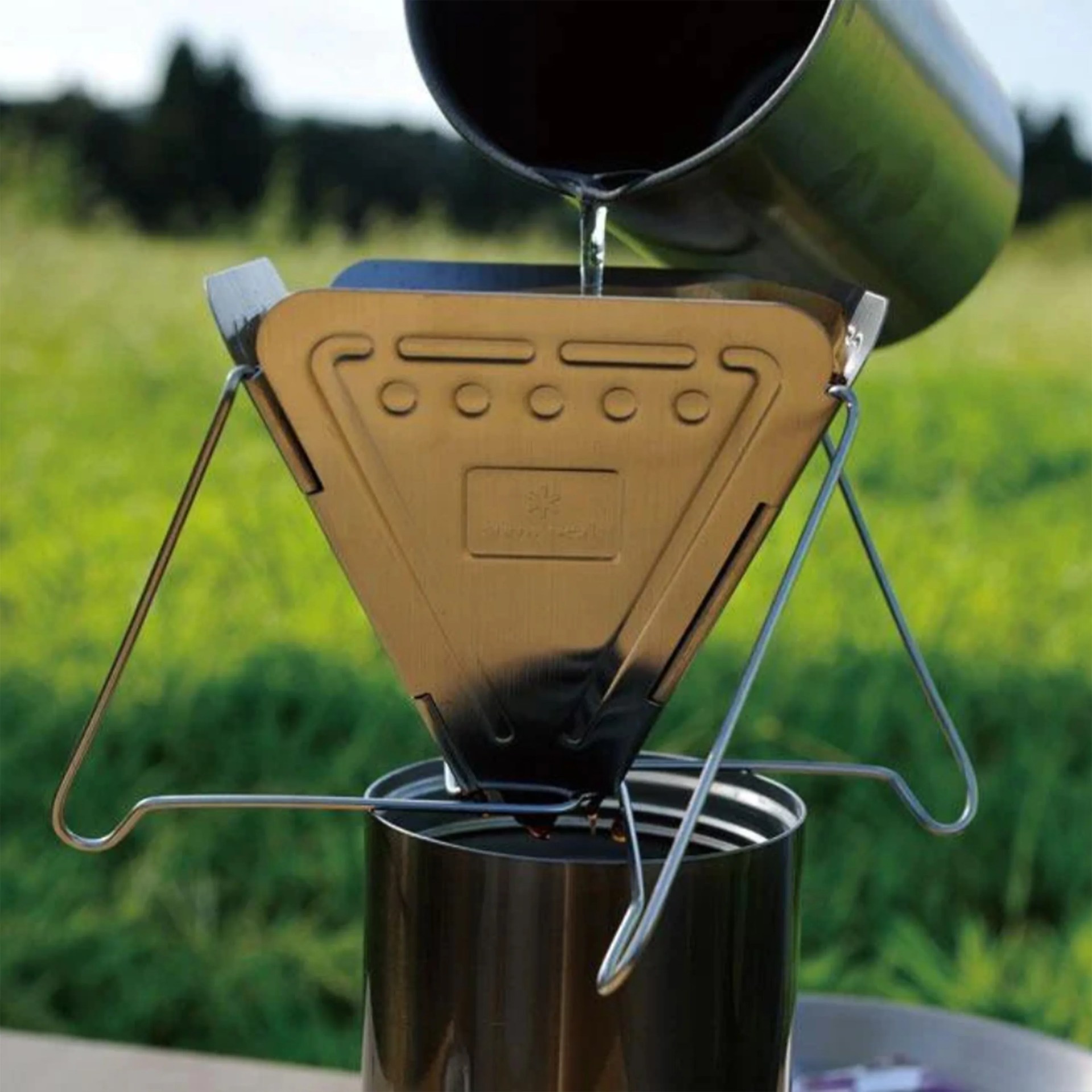 Metallic triangular coffee dripper with wire stand placed on a metal cup outdoors with water being poured into it.