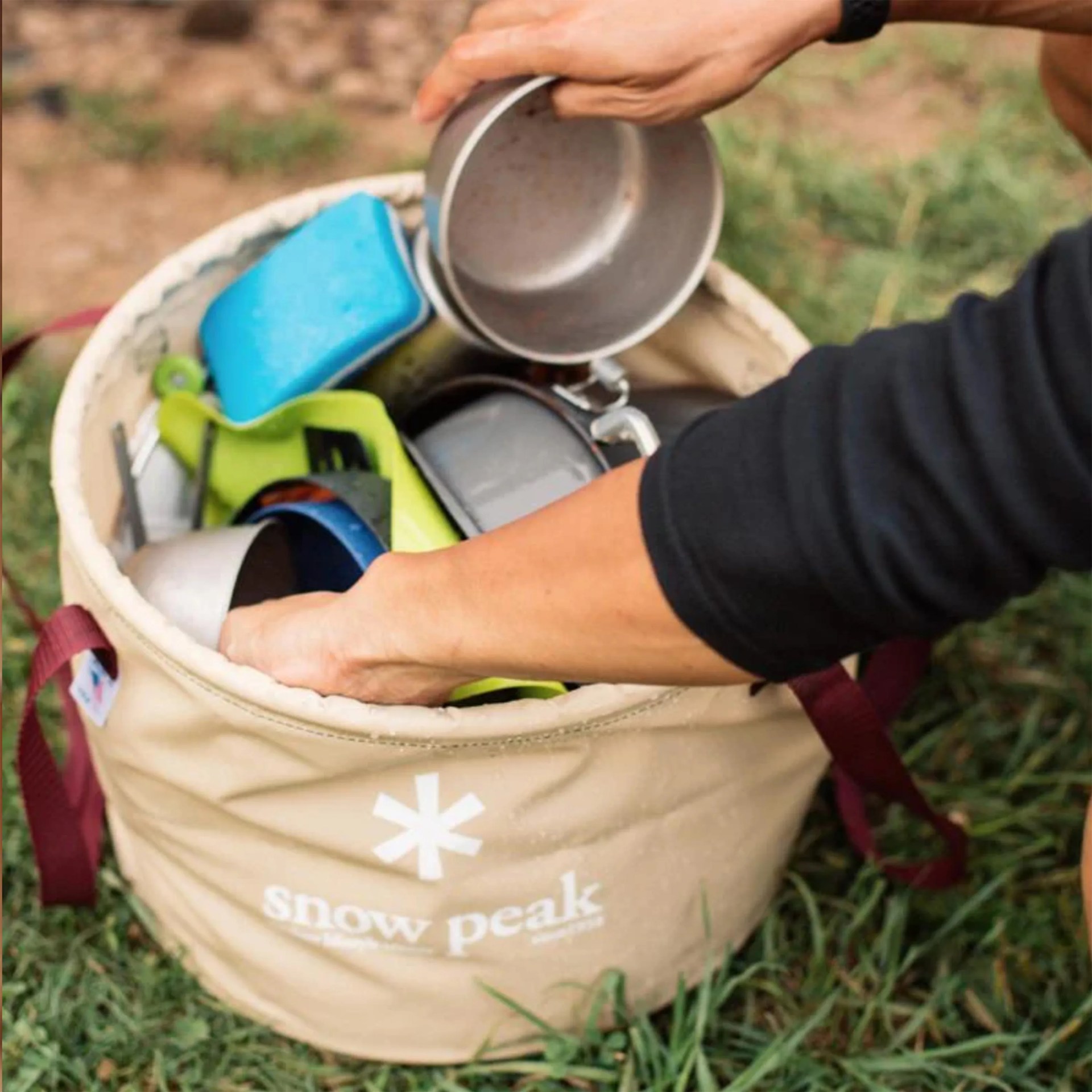 Beige Snow Peak canvas bucket filled with camping cookware and a person&rsquo;s hands holding a metal cup.