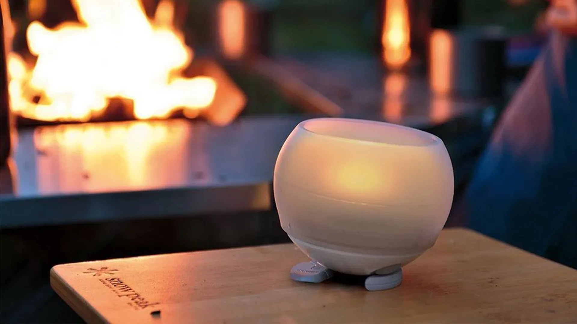 Small round white candle holder with a lit candle inside on a wooden table near a fire.