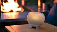 Small round white candle holder with a lit candle inside on a wooden table near a fire.