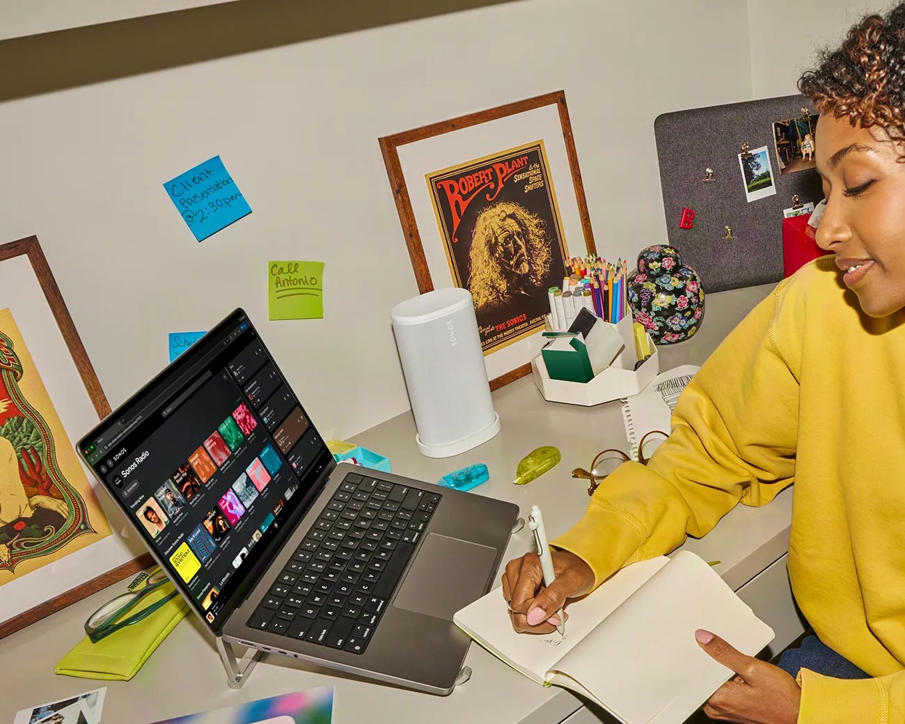 Person in a yellow sweatshirt writing in a notebook at a desk with a laptop, white Sonos speaker, and framed posters.