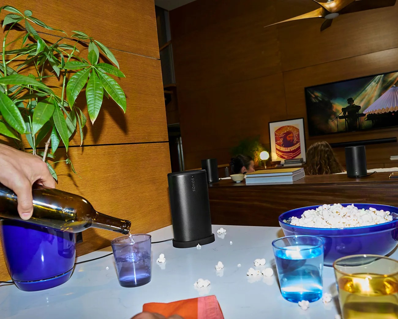 Blue plastic bowl filled with popcorn and a hand pouring water from a brown bottle into a purple glass on a white table.