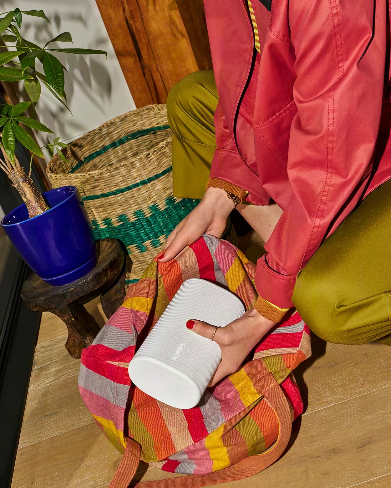 White Sonos speaker being placed into a colorful striped fabric bag by a person wearing a red jacket and olive green pants.
