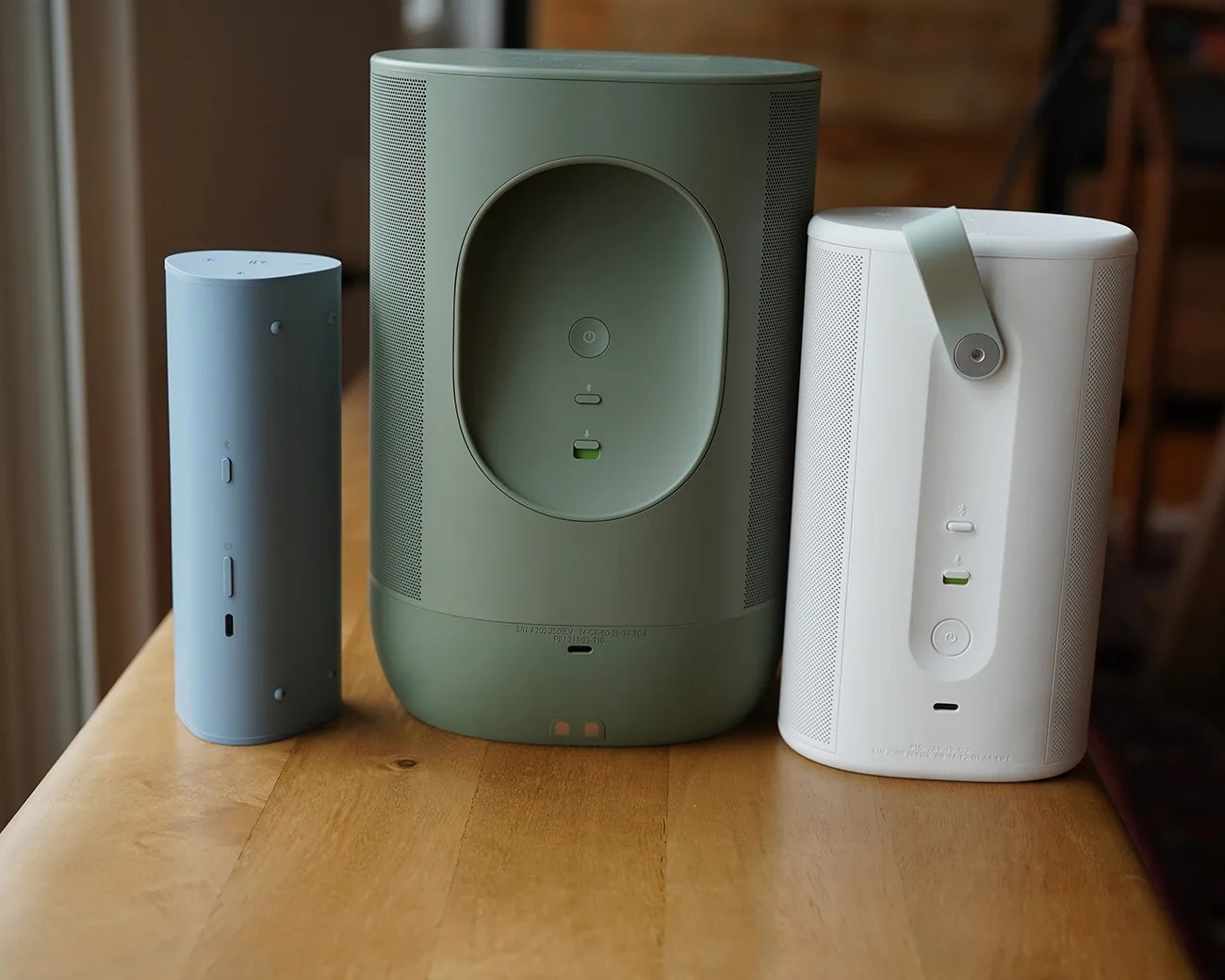 Three cylindrical wireless speakers in blue, green, and white, placed side by side on a wooden table.