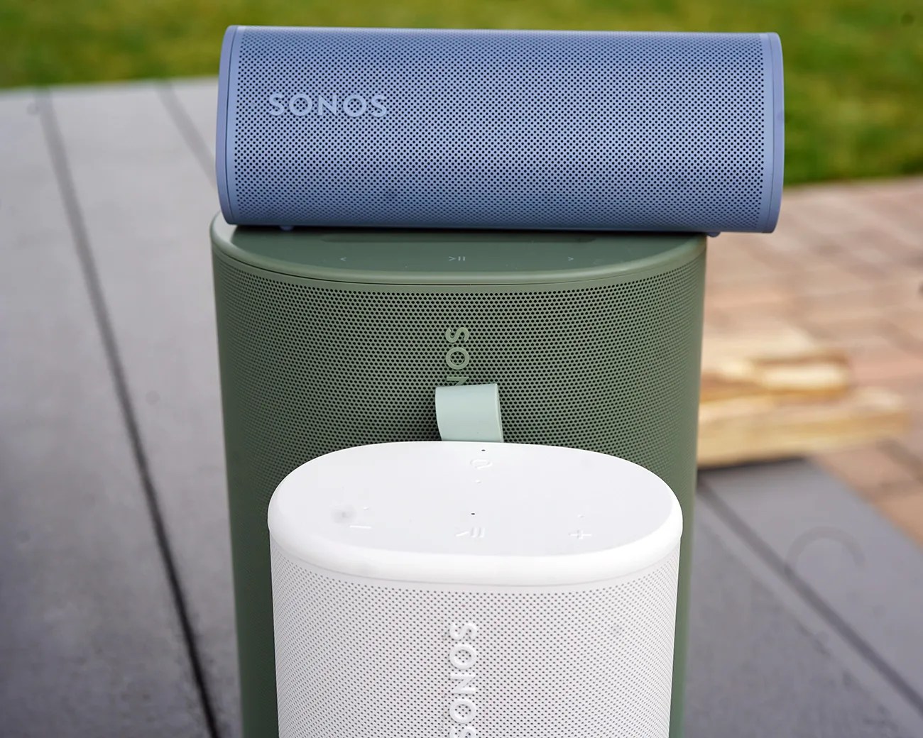 Three Sonos speakers stacked outdoors: a blue cylindrical speaker on top, a large green cylindrical speaker in the middle, and a smaller white oval speaker at the bottom.