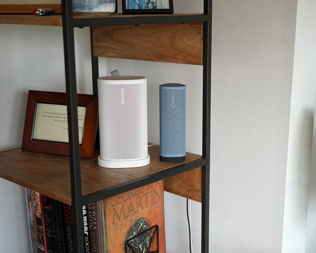 Two Sonos speakers, one white and one blue, placed on a wooden shelf with a black metal frame.