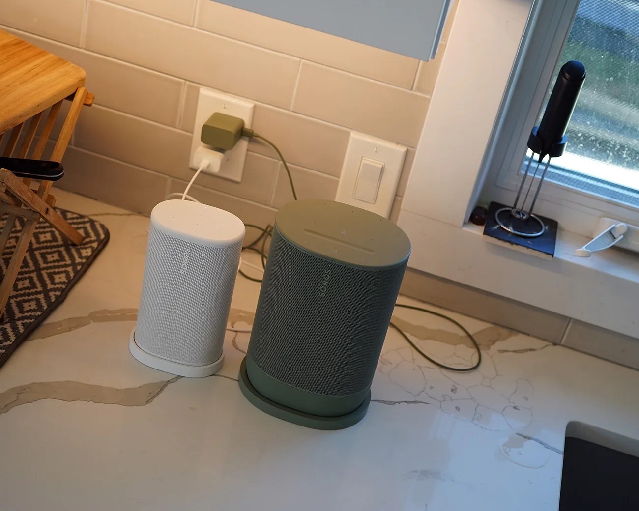 Two Sonos speakers, one white and one green, placed on a kitchen countertop near a window and electrical outlet.