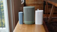 Three Sonos wireless speakers in blue, green, and white on a wooden table near a window.