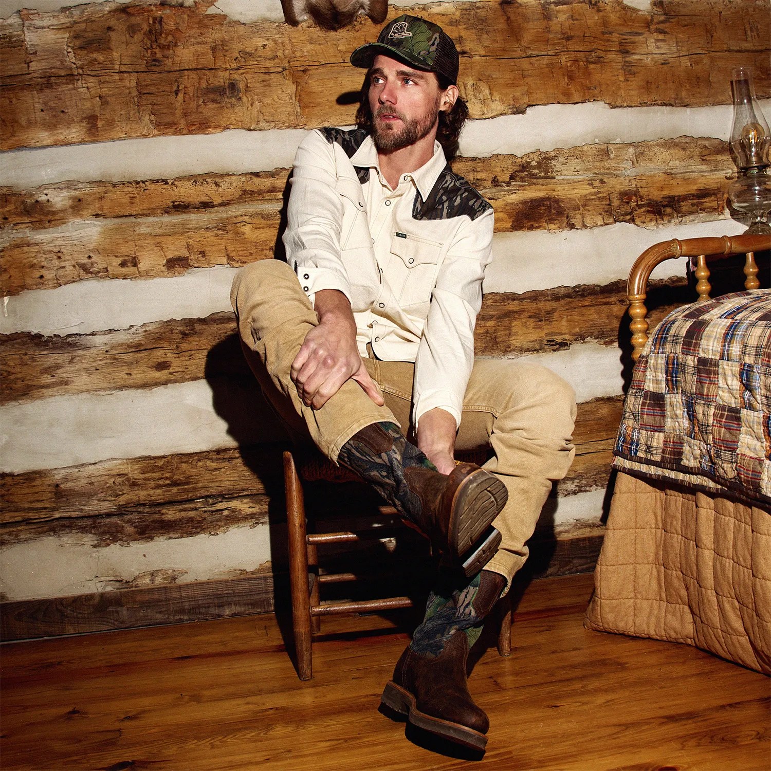 Man wearing a cream shirt with dark shoulder patches, tan pants, and brown cowboy boots sitting on a wooden chair in a log cabin.
