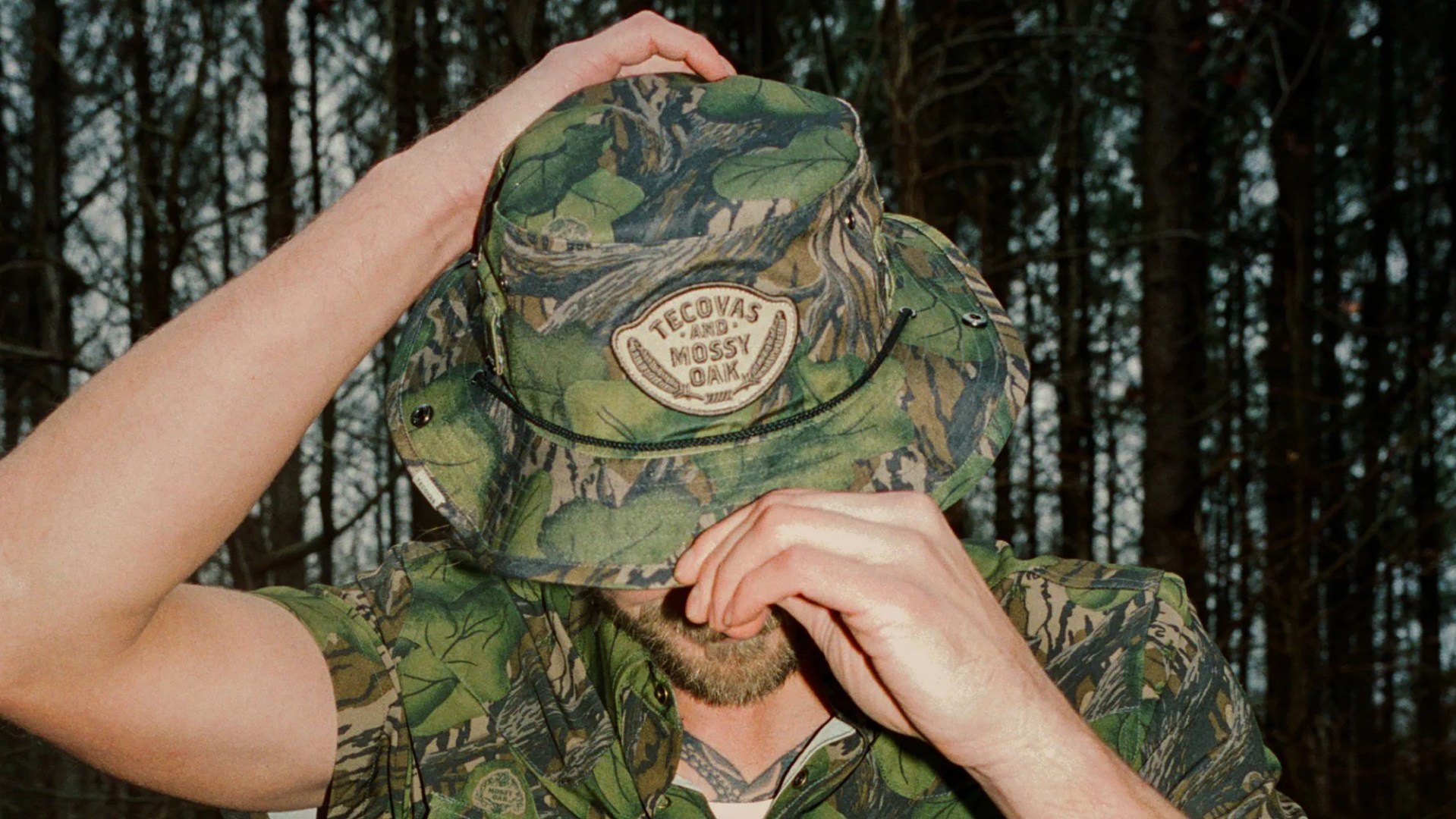 Green and brown camouflage bucket hat with "Tecovas and Mossy Oak" patch worn by a person in a forest.
