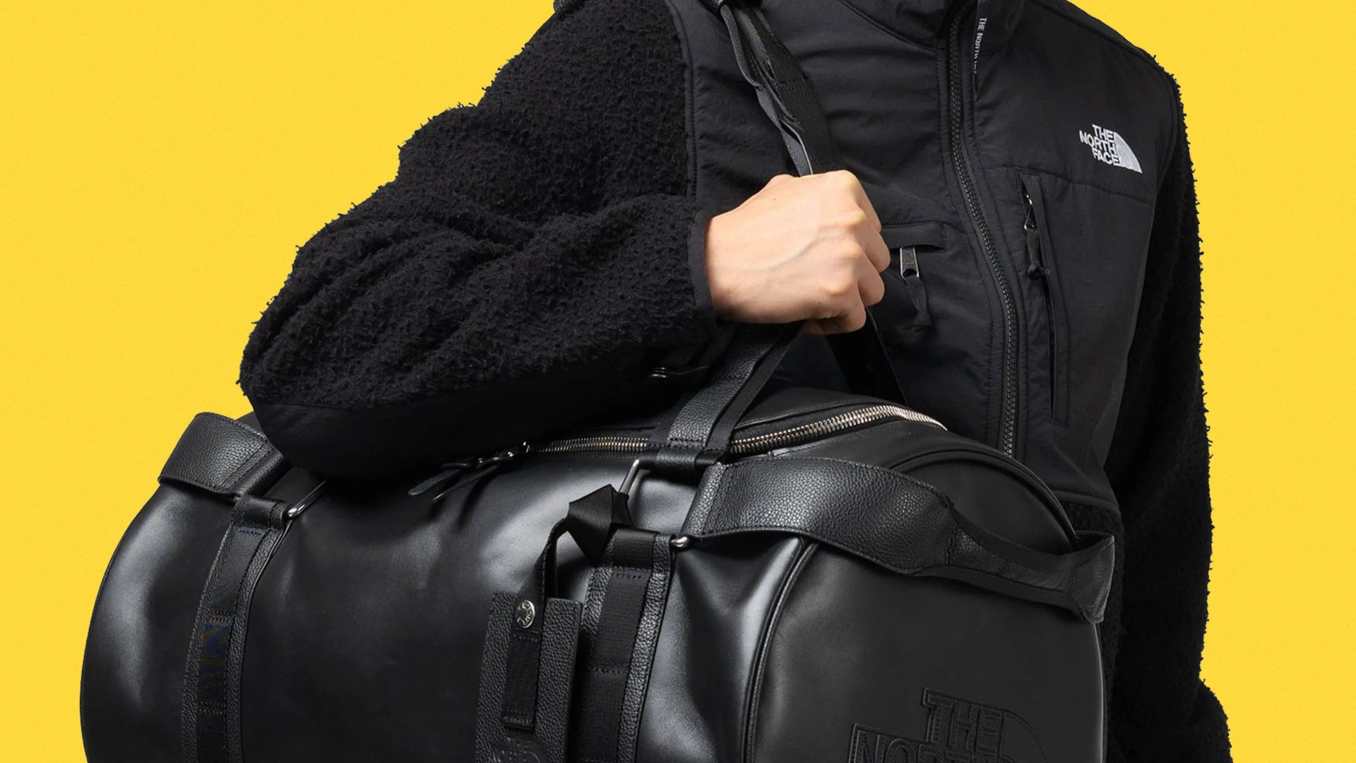 Black leather duffel bag with embossed logo held by a person wearing a black fleece jacket with a white logo.