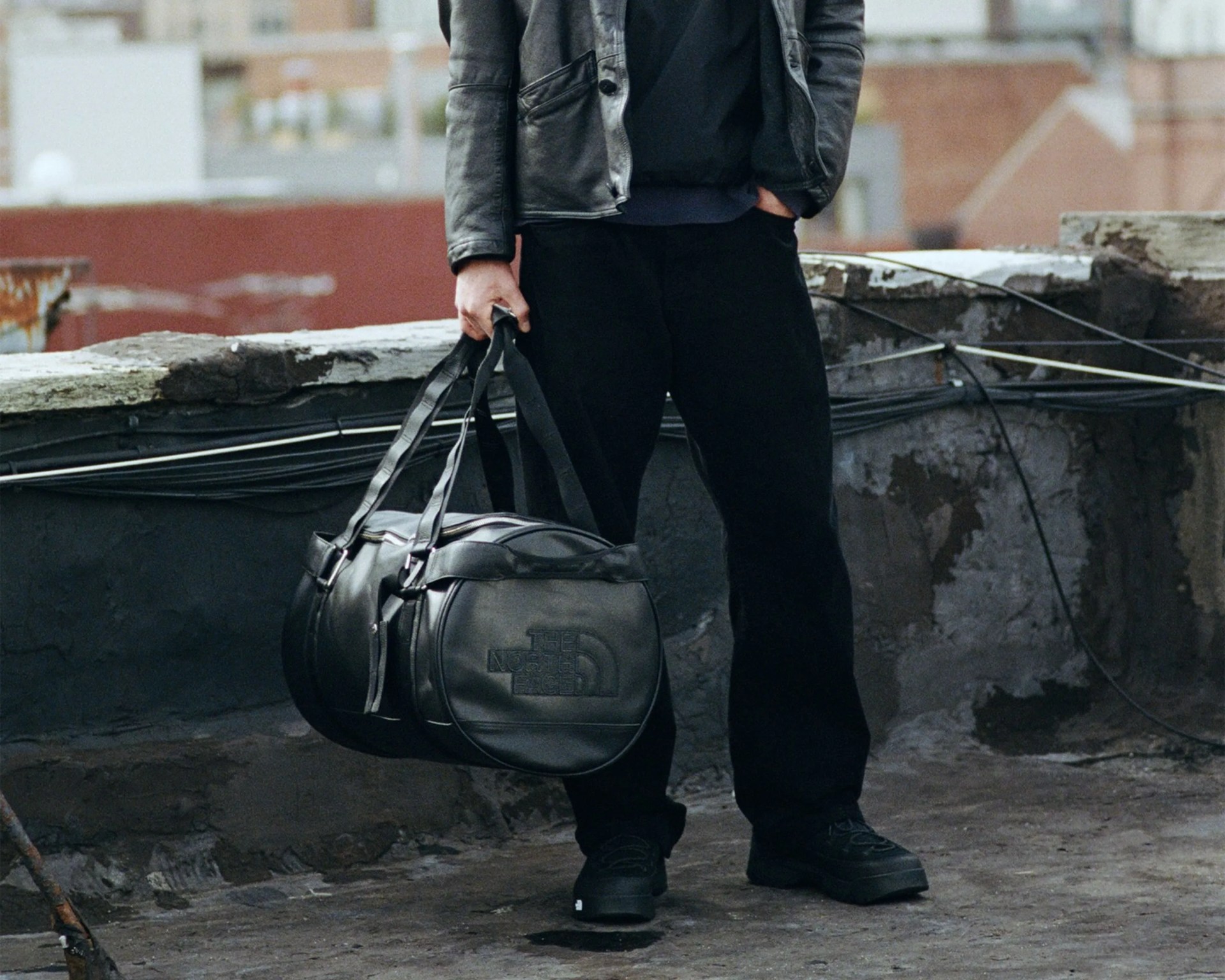 Person holding a black leather The North Face duffel bag, wearing black pants and shoes, standing on a rooftop.