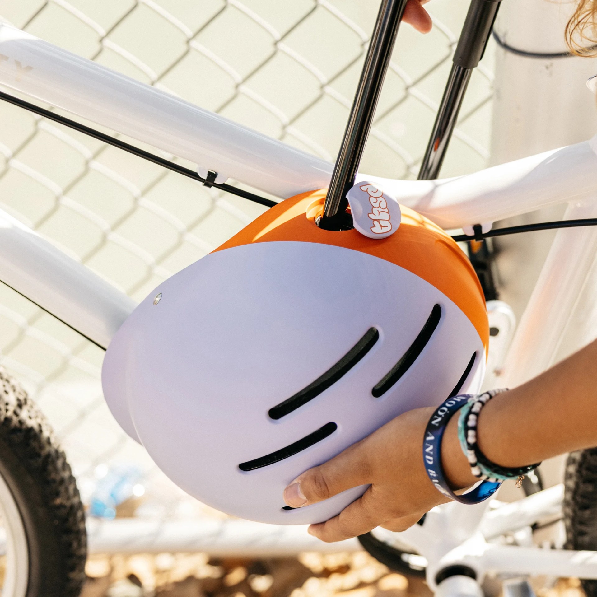 Orange and white bike helmet being held near a white bicycle frame with a chain-link fence in the background.