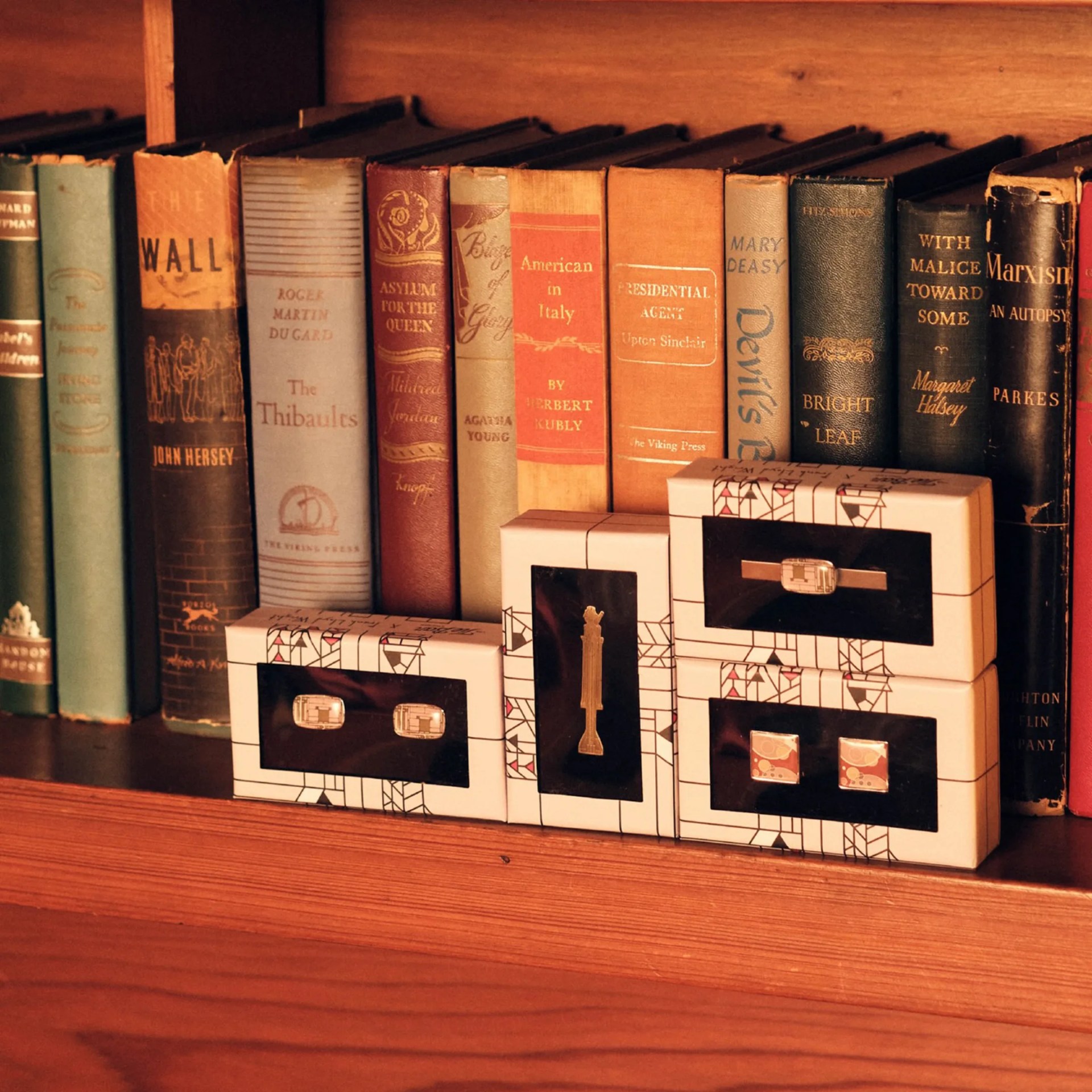 Four sets of cufflinks in white and black geometric patterned boxes on a wooden shelf in front of vintage books.