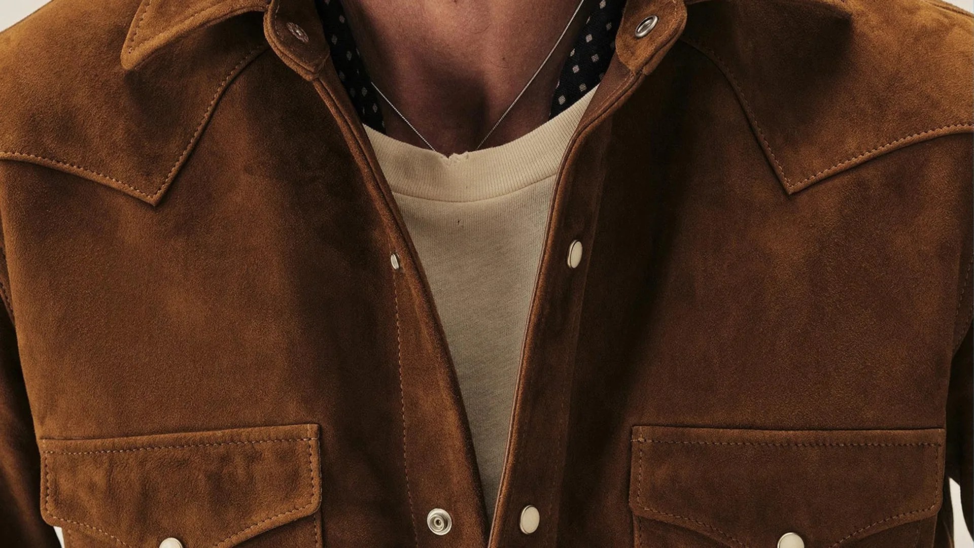 Brown suede jacket with snap buttons and flap pockets worn over a cream crewneck shirt.
