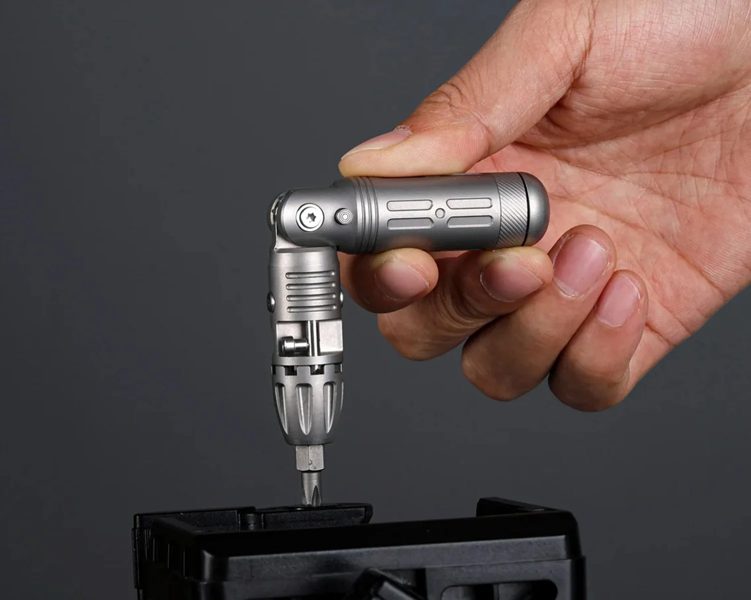 Compact silver multi-angle screwdriver held in a hand, tightening a screw on a black plastic object.