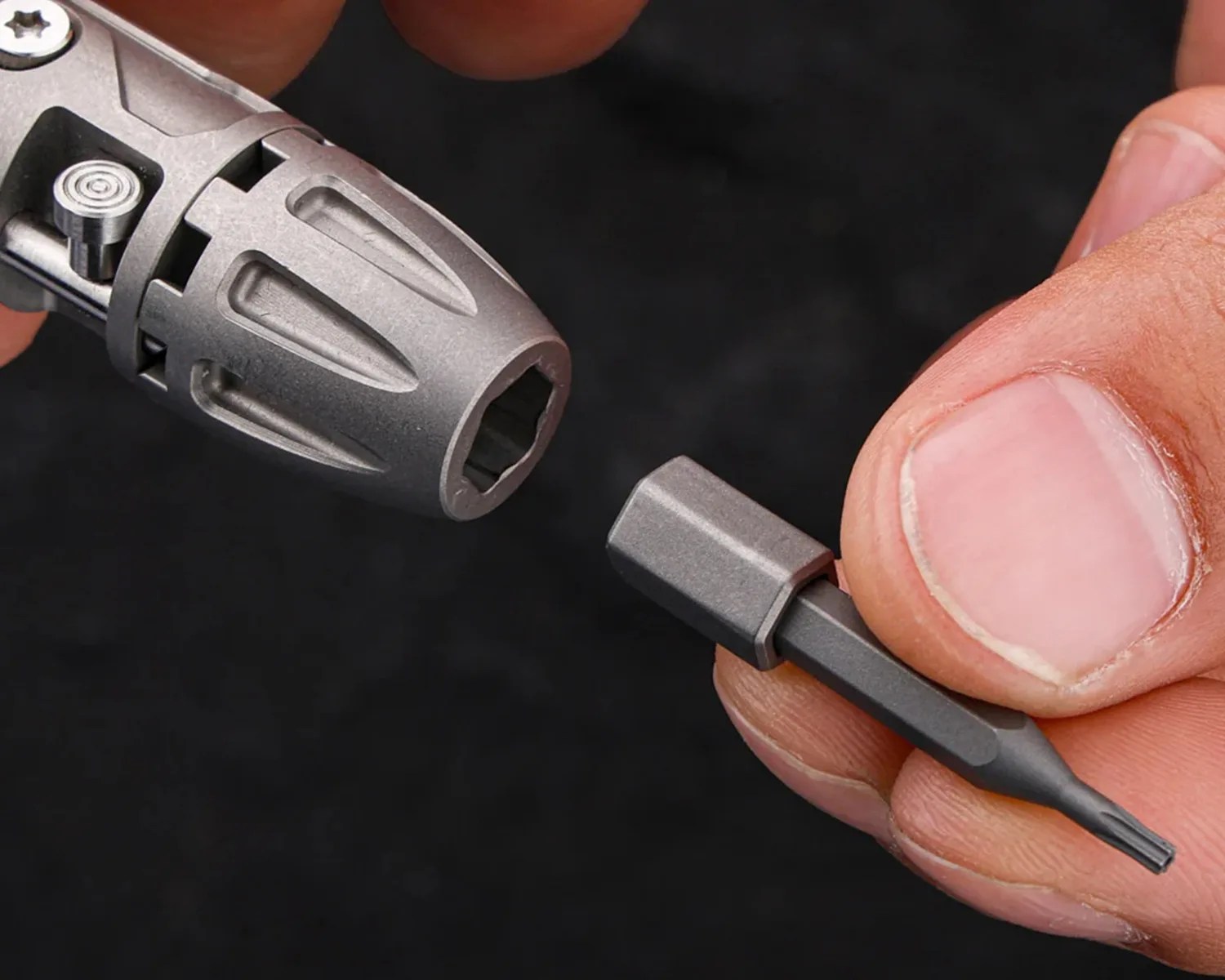 Close-up of a hand holding a small metal screwdriver bit near a metallic tool chuck against a black background.