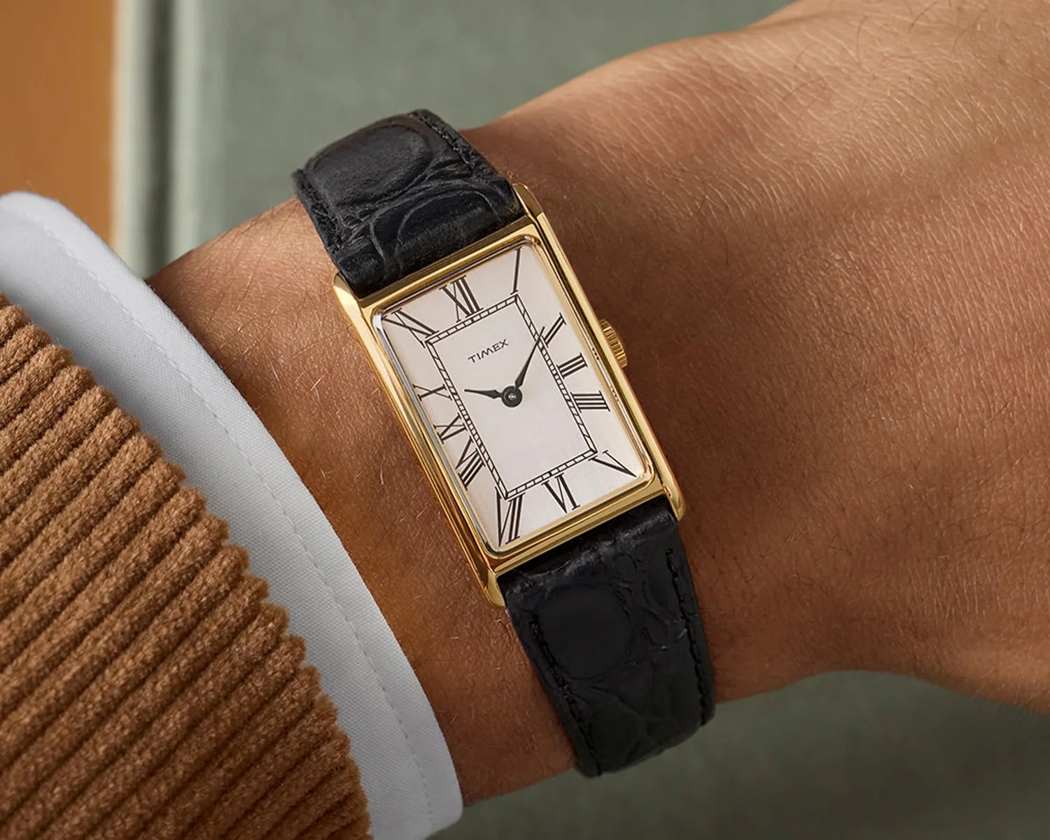 Rectangular gold-tone Timex wristwatch with white dial, black Roman numerals, and black leather strap worn on a wrist.
