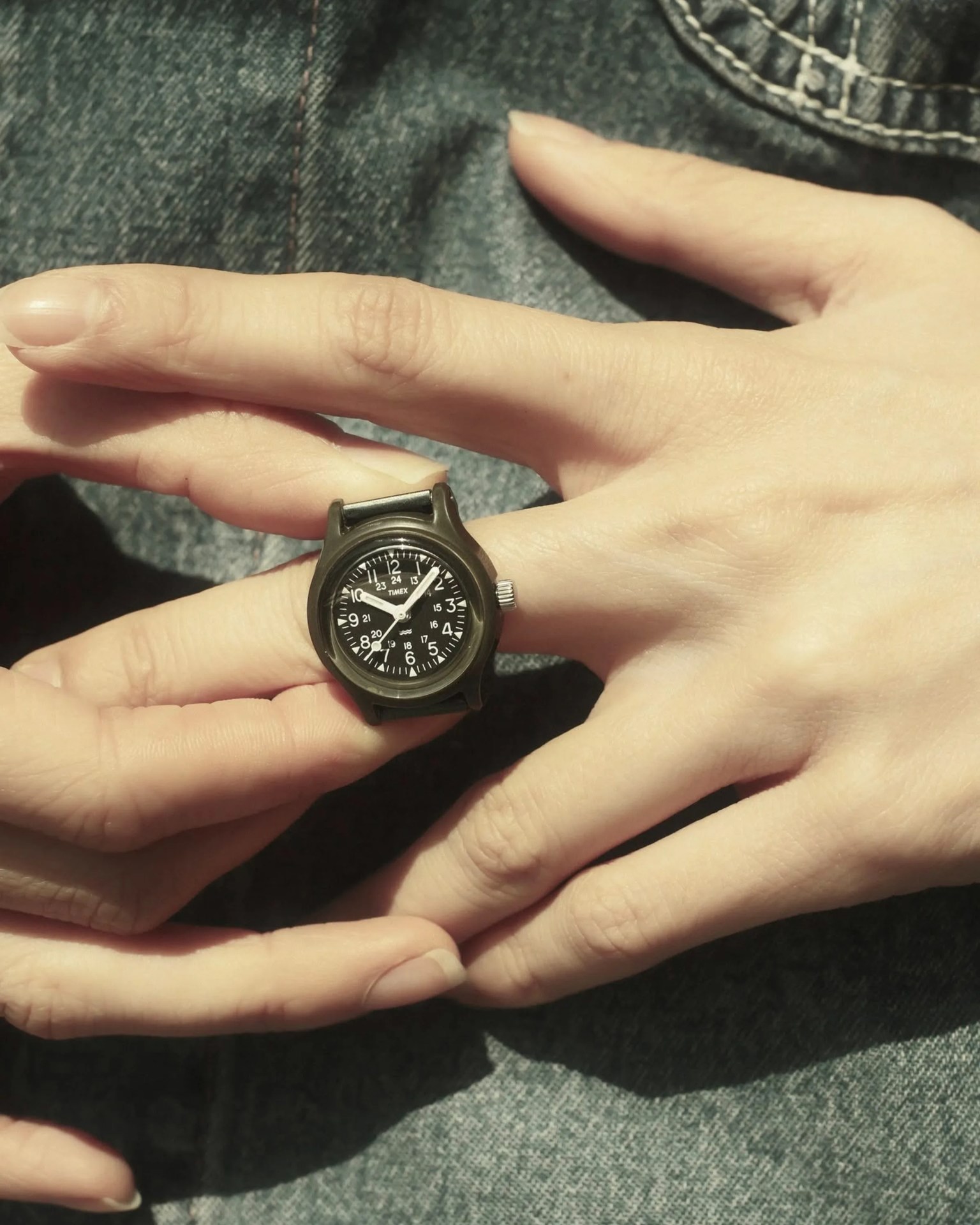 Black Timex watch face with white numerals held between fingers over denim fabric.