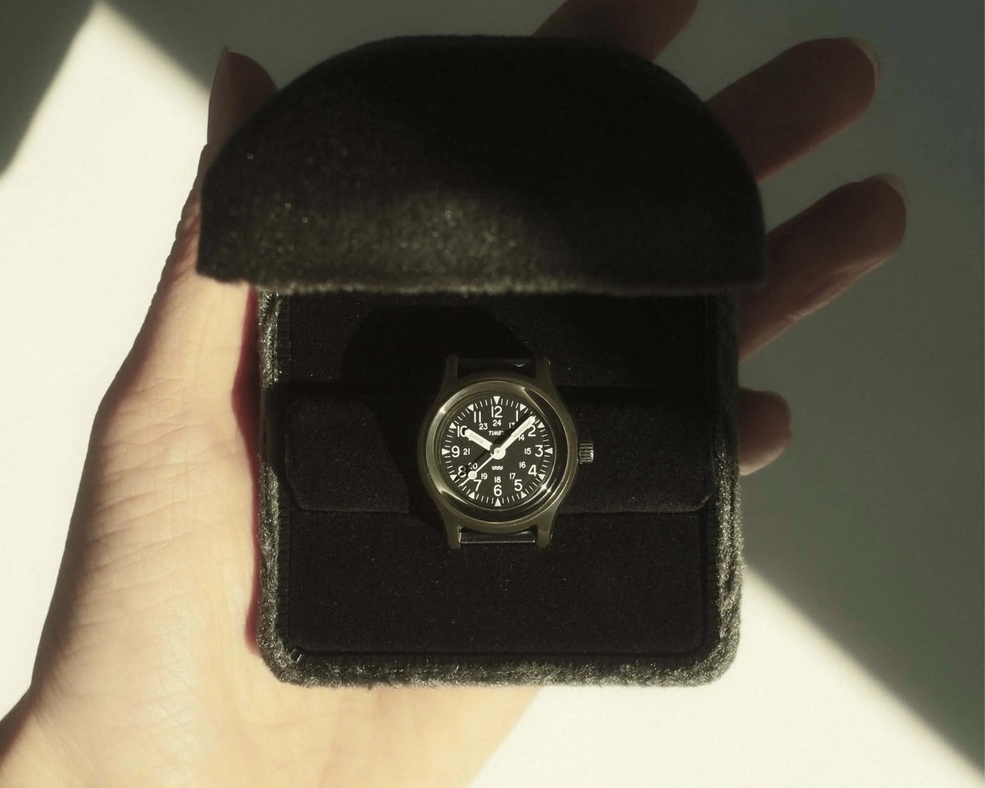 Black Timex wristwatch with white numerals and hands in a black fabric case held in a hand.
