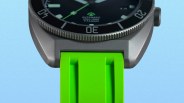 Close-up of a titanium watch case with a black bezel and bright green rubber strap against a blue background.