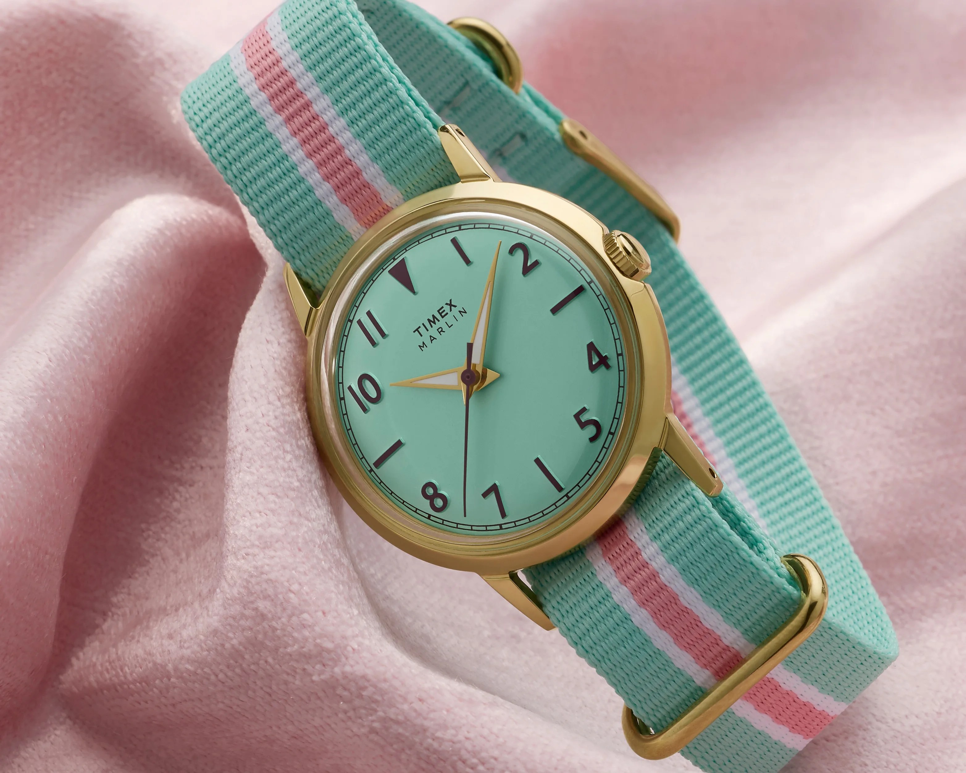 Gold Timex Marlin watch with mint green dial and striped mint, white, and pink fabric strap on pink fabric.