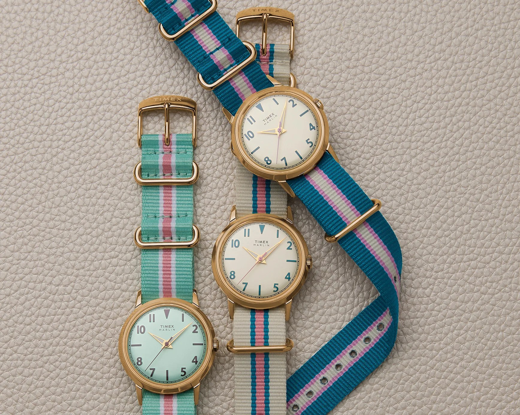 Three Timex Marlin watches with gold cases and striped fabric straps in teal, beige, and turquoise on a textured beige surface.