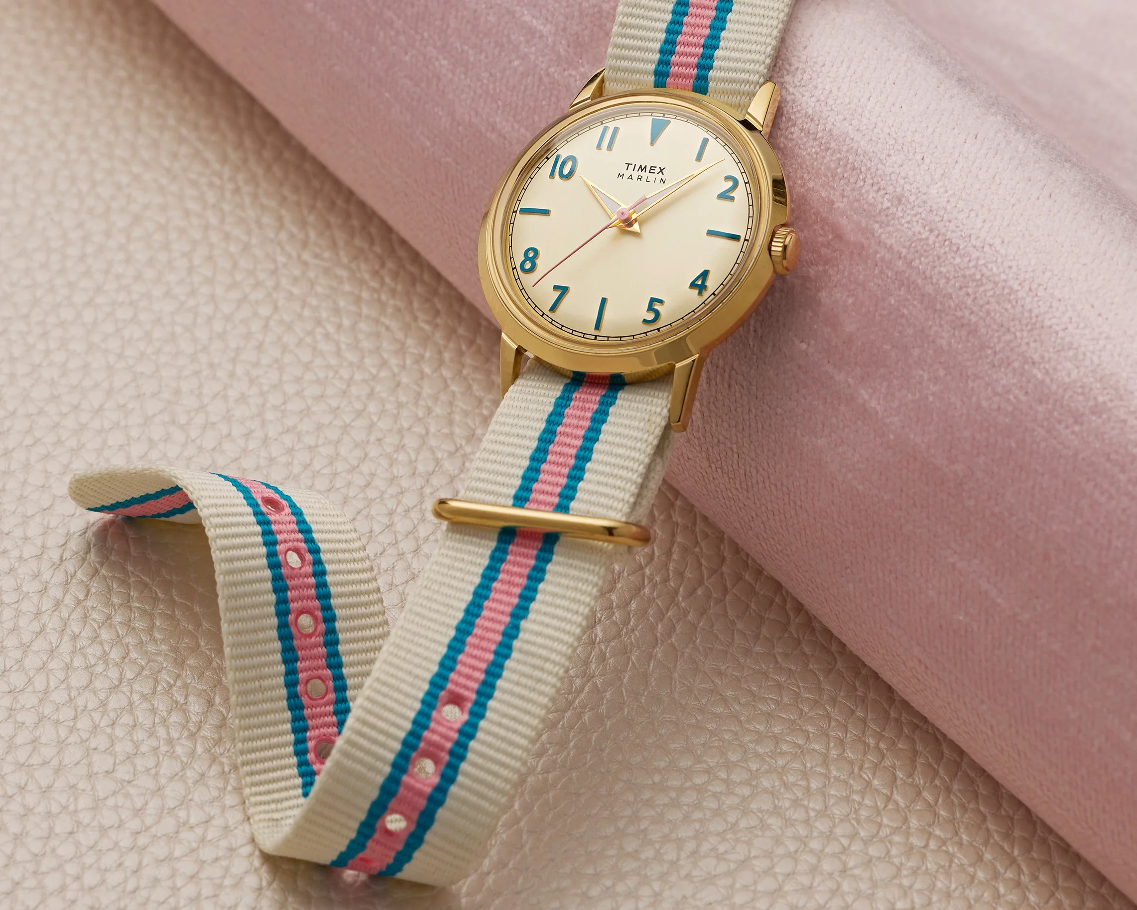Gold Timex Marlin watch with cream dial and blue numerals on a cream fabric strap with pink and blue stripes.