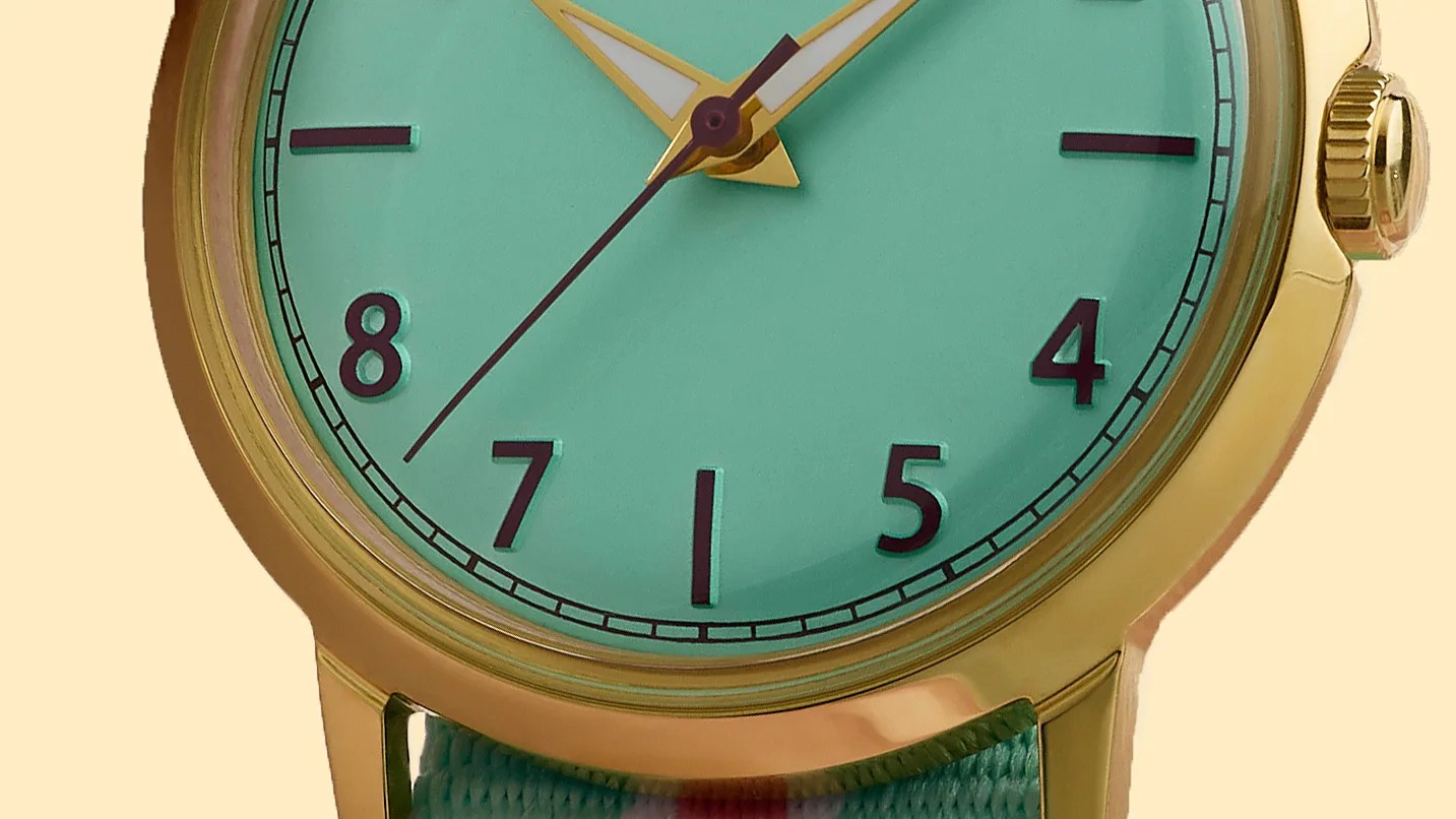 Close-up of a gold watch with a turquoise face, black numerals, and a matching turquoise fabric strap.