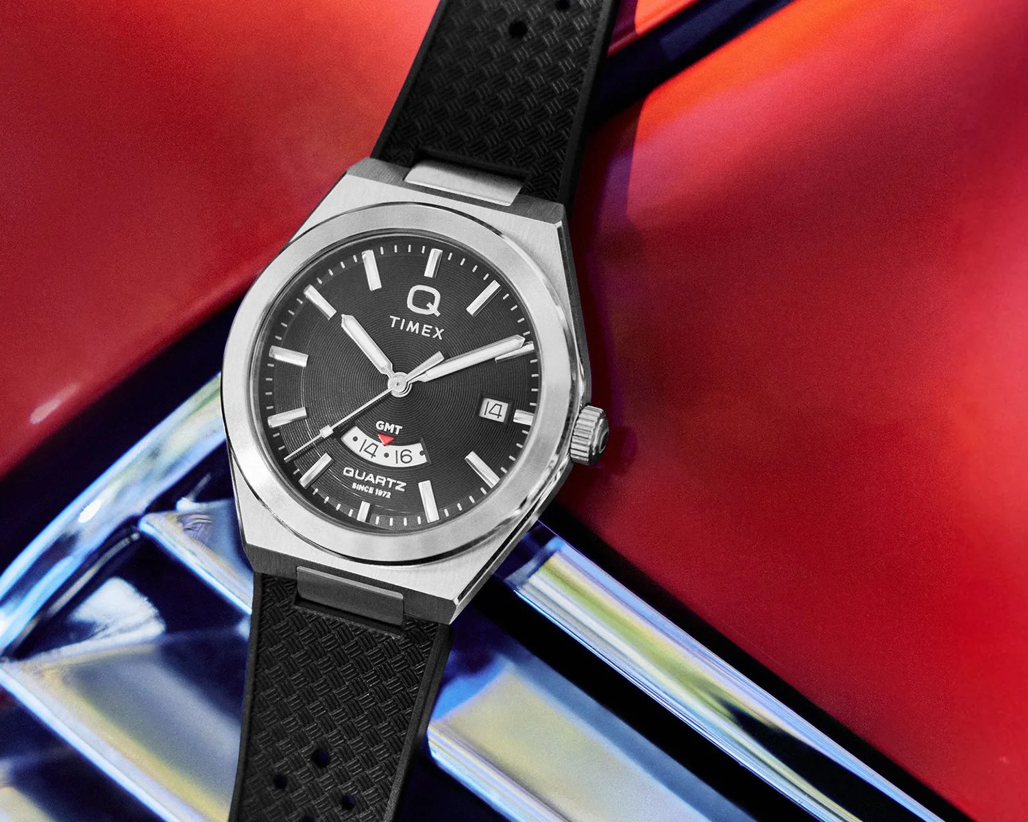 Timex Q Quartz wristwatch with black textured dial, date window, and black patterned rubber strap on a red and chrome surface.