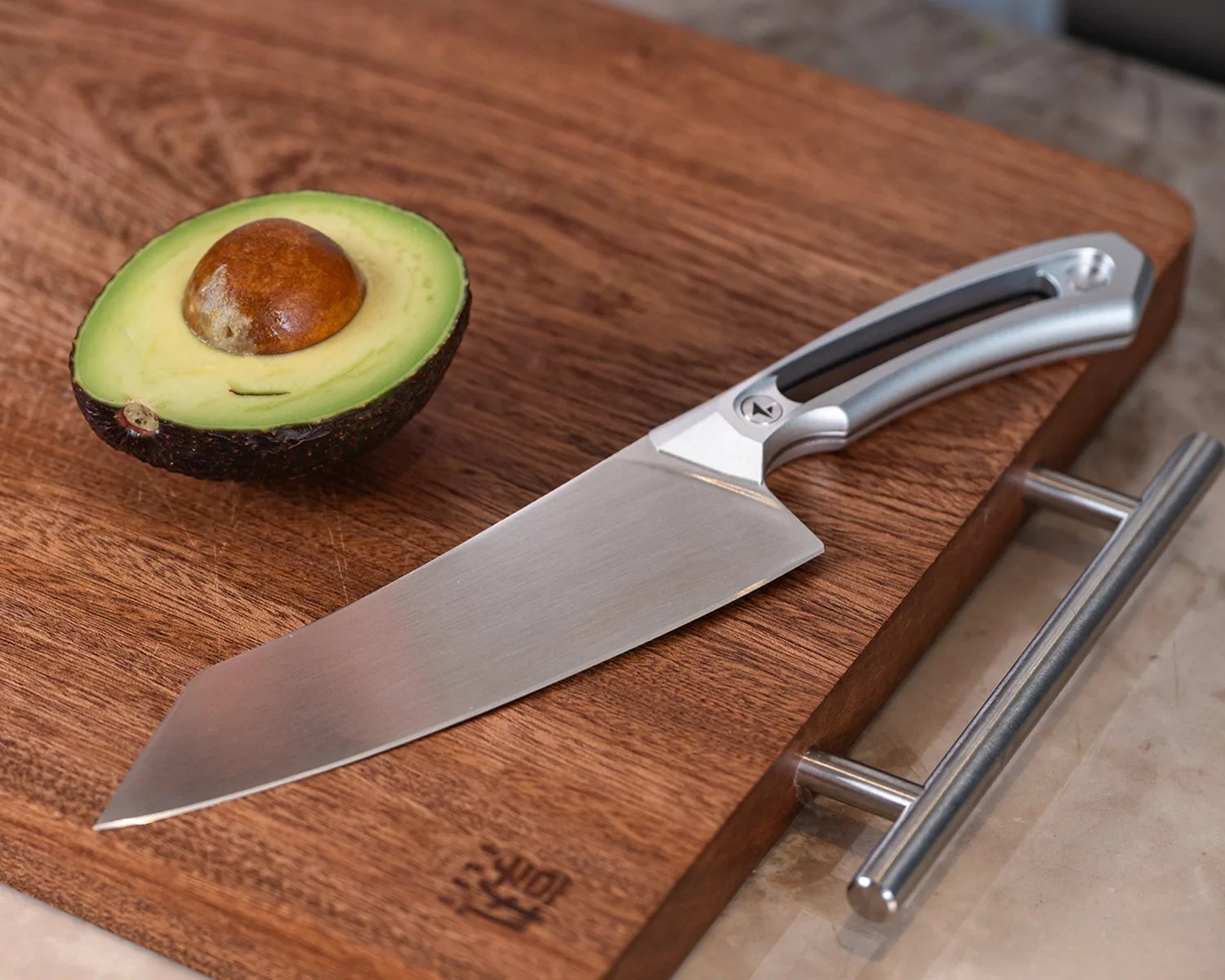 Silver kitchen knife with a black and silver handle on a wooden cutting board next to a halved avocado with the pit.
