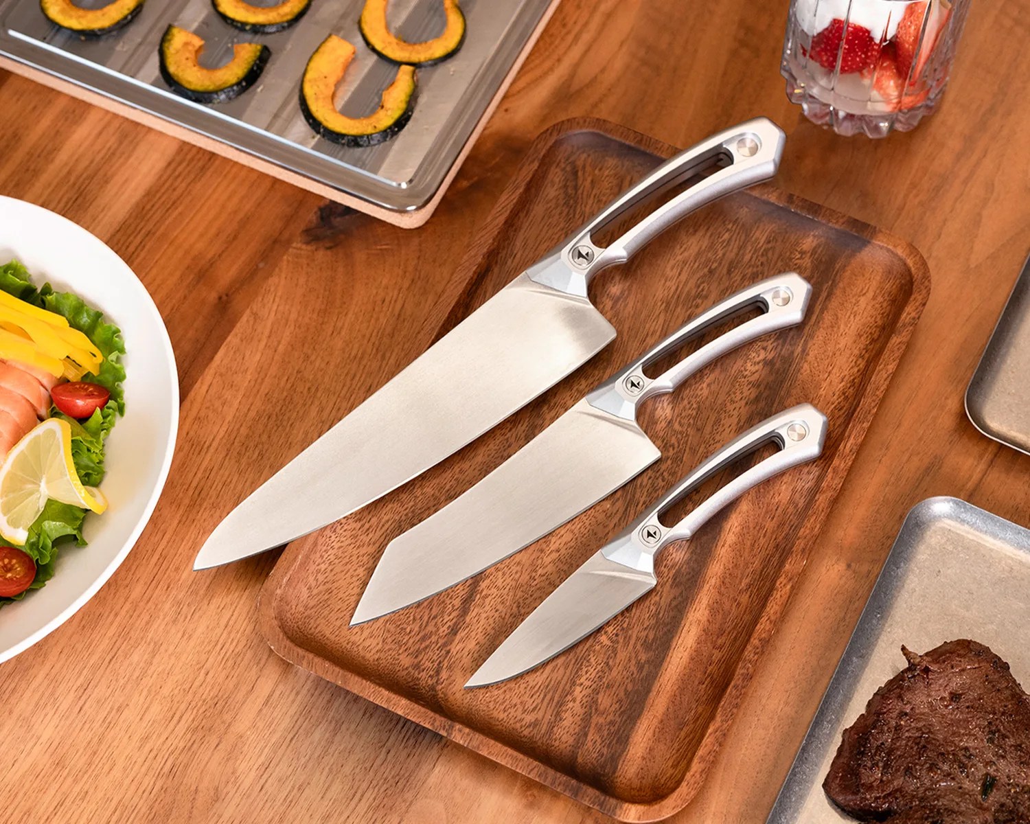 Three stainless steel kitchen knives with silver handles on a wooden tray, surrounded by food items on a wooden table.