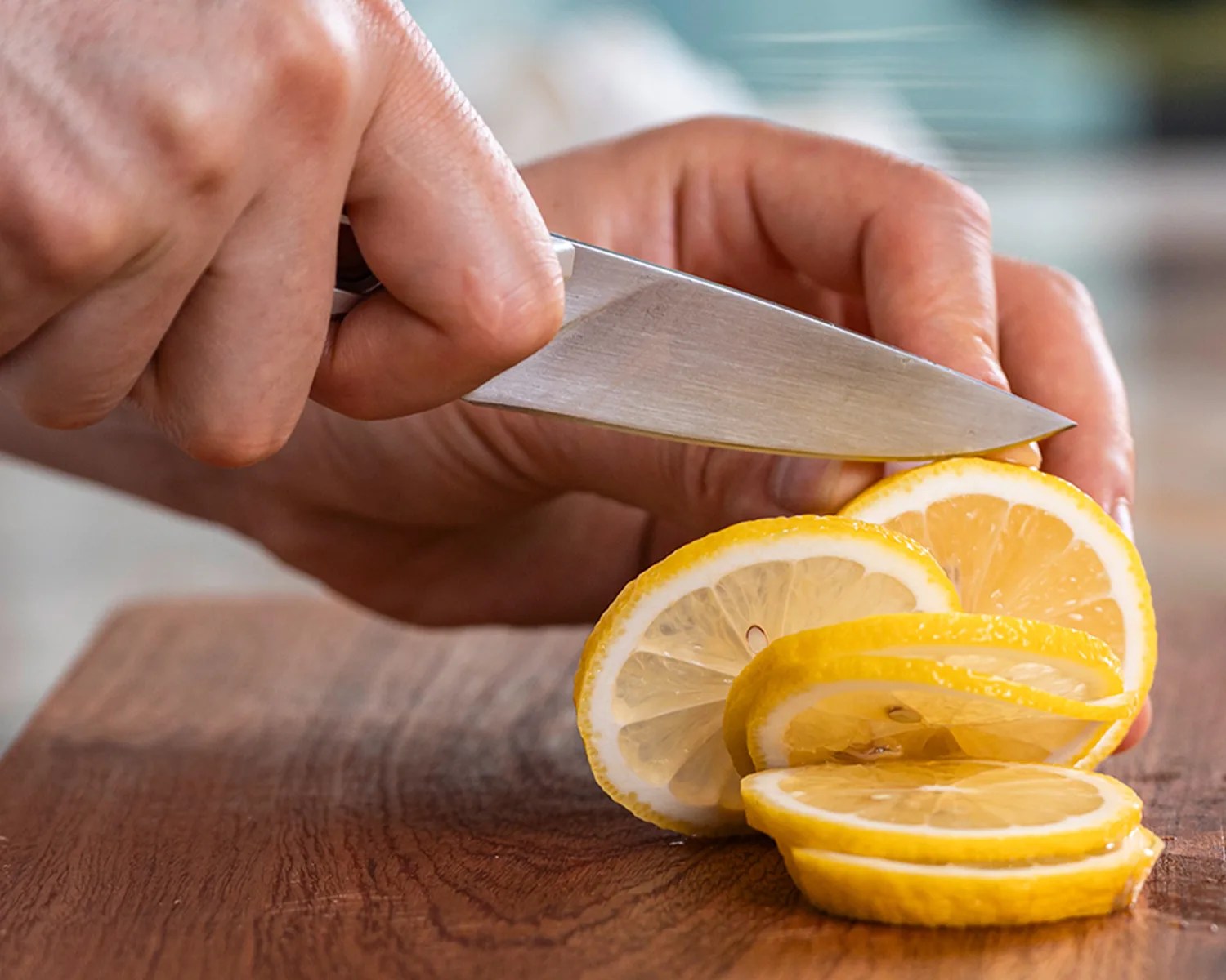 Hand holding a knife slicing a lemon into thin rounds on a wooden cutting board.