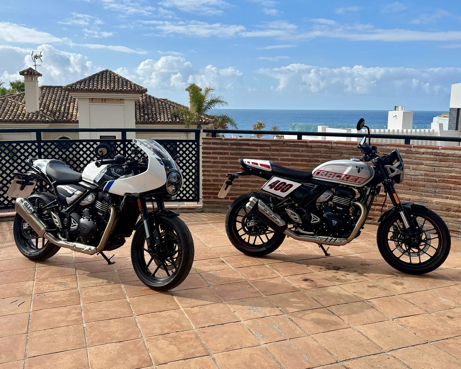 Two white and black Triumph motorcycles parked on a tiled terrace with ocean and houses in the background.