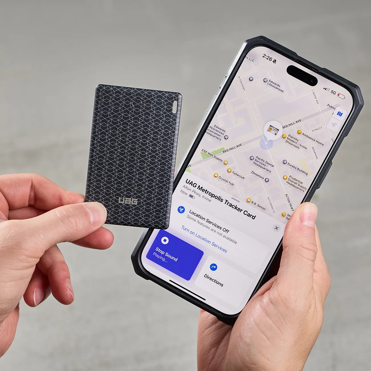 Hand holding a black UAG Metropolis Tracker Card next to a smartphone displaying its location on a map.