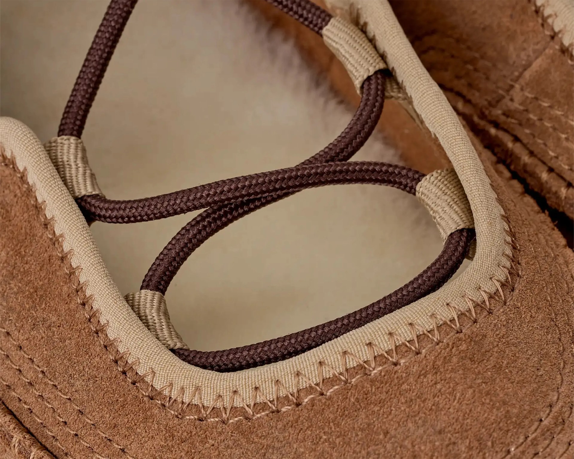 Close-up of brown suede shoe with beige trim and dark brown round laces threaded through fabric loops.
