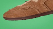 Brown suede moccasin shoe with detailed stitching and a dark brown rubber sole on a green background.