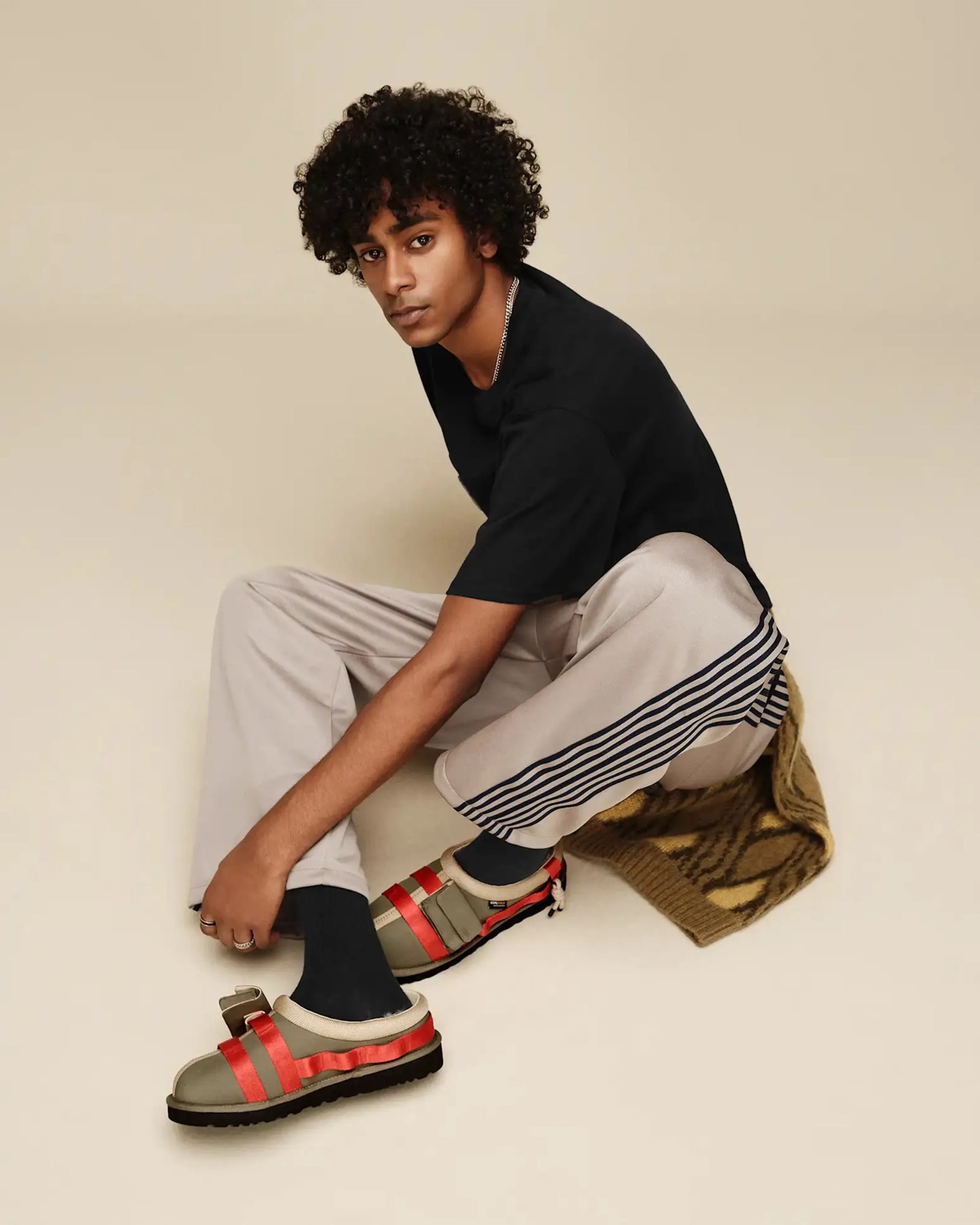 Person wearing beige pants with black stripes, black socks, olive green sandals with red straps, and a black t-shirt sitting on a beige floor.