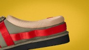 Close-up of an olive green shoe with beige padded collar, red strap detail, and black sole against a yellow background.
