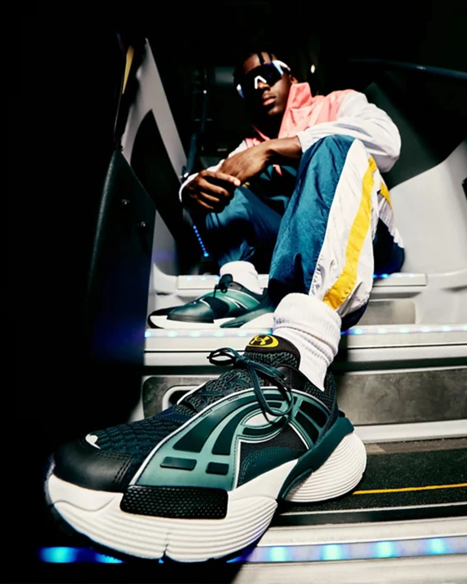 Black and teal Under Armour sneakers with white soles worn by a person in blue and yellow track pants sitting on stairs.