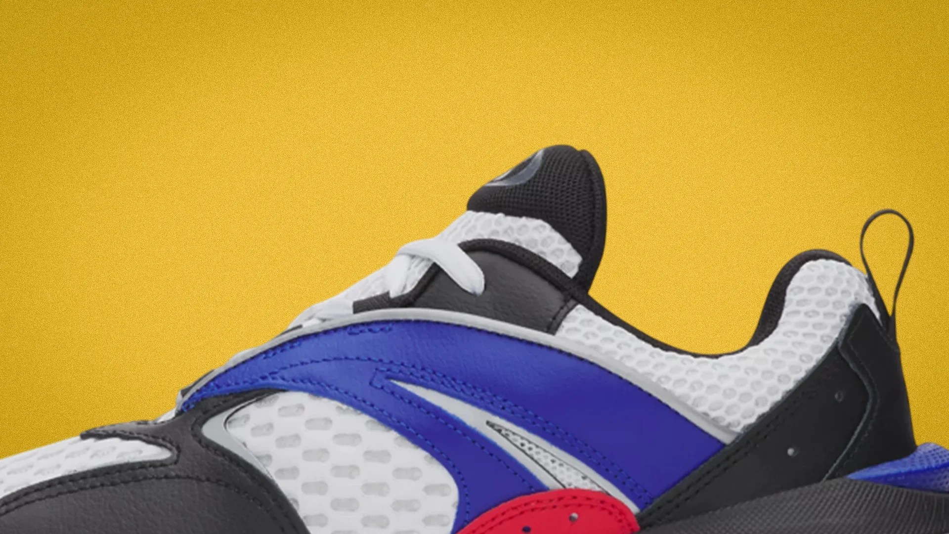 Close-up of a white mesh sneaker with black, blue, and red leather overlays against a yellow background.