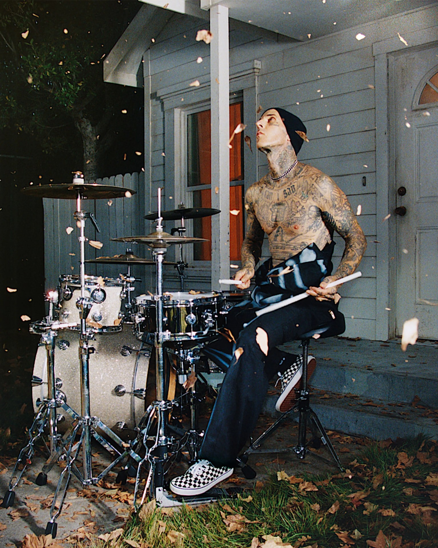 Tattooed person wearing a beanie and checkered slip-on shoes playing a drum set outdoors with leaves falling.