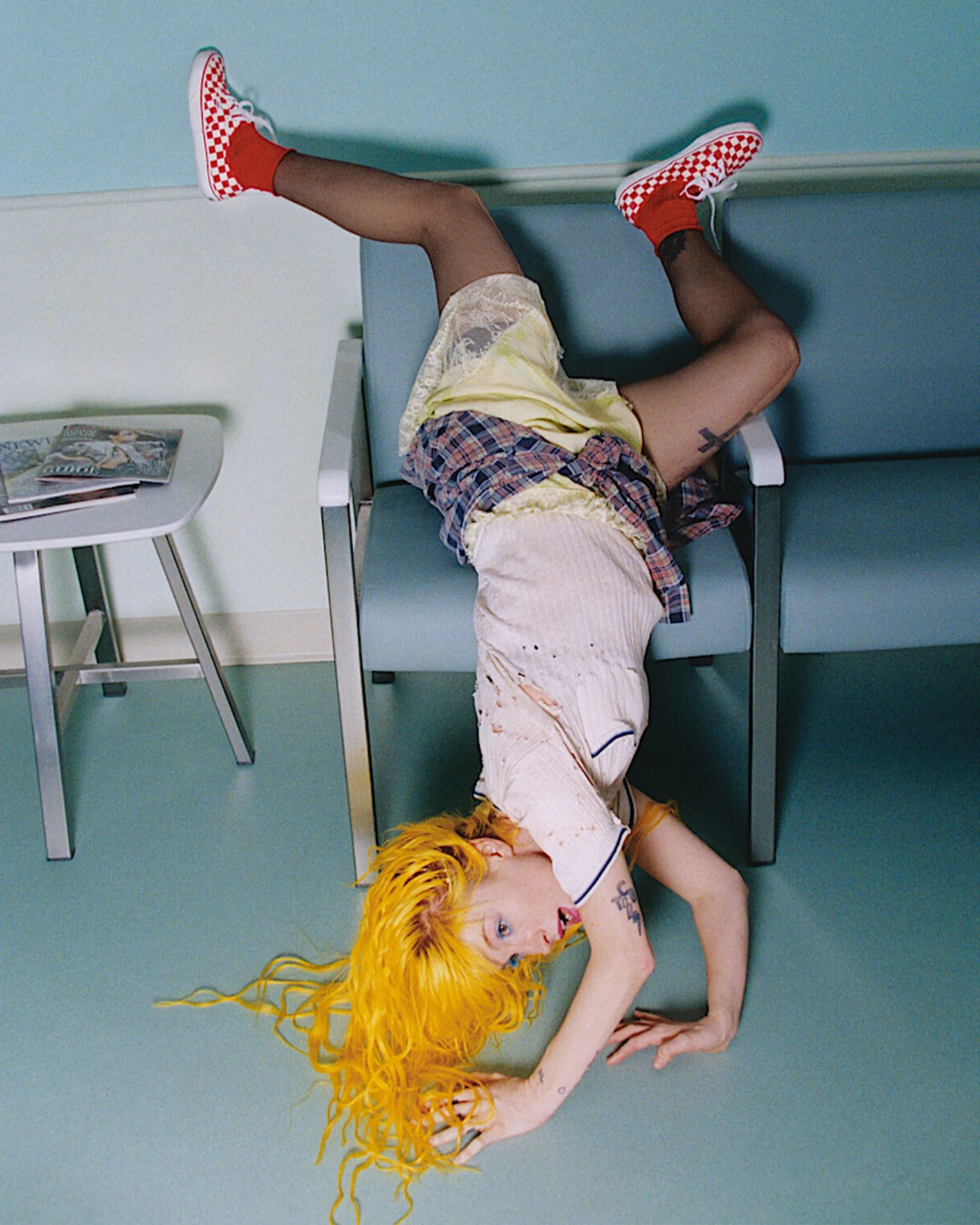 Person with long bright yellow hair wearing a stained white shirt, yellow slip, plaid shirt, and red checkered sneakers upside down on a blue chair.