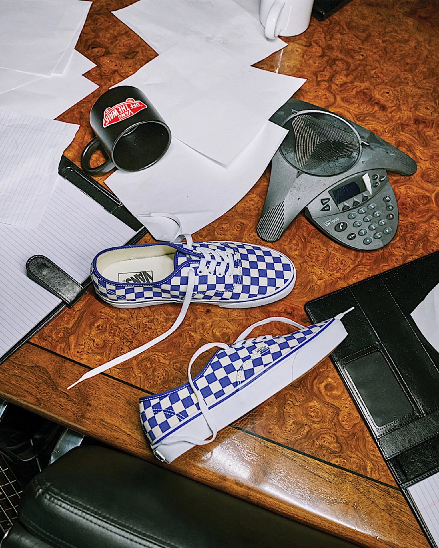 Pair of blue and white checkered Vans sneakers with white laces on a wooden office desk with papers, a black mug, and a conference phone.