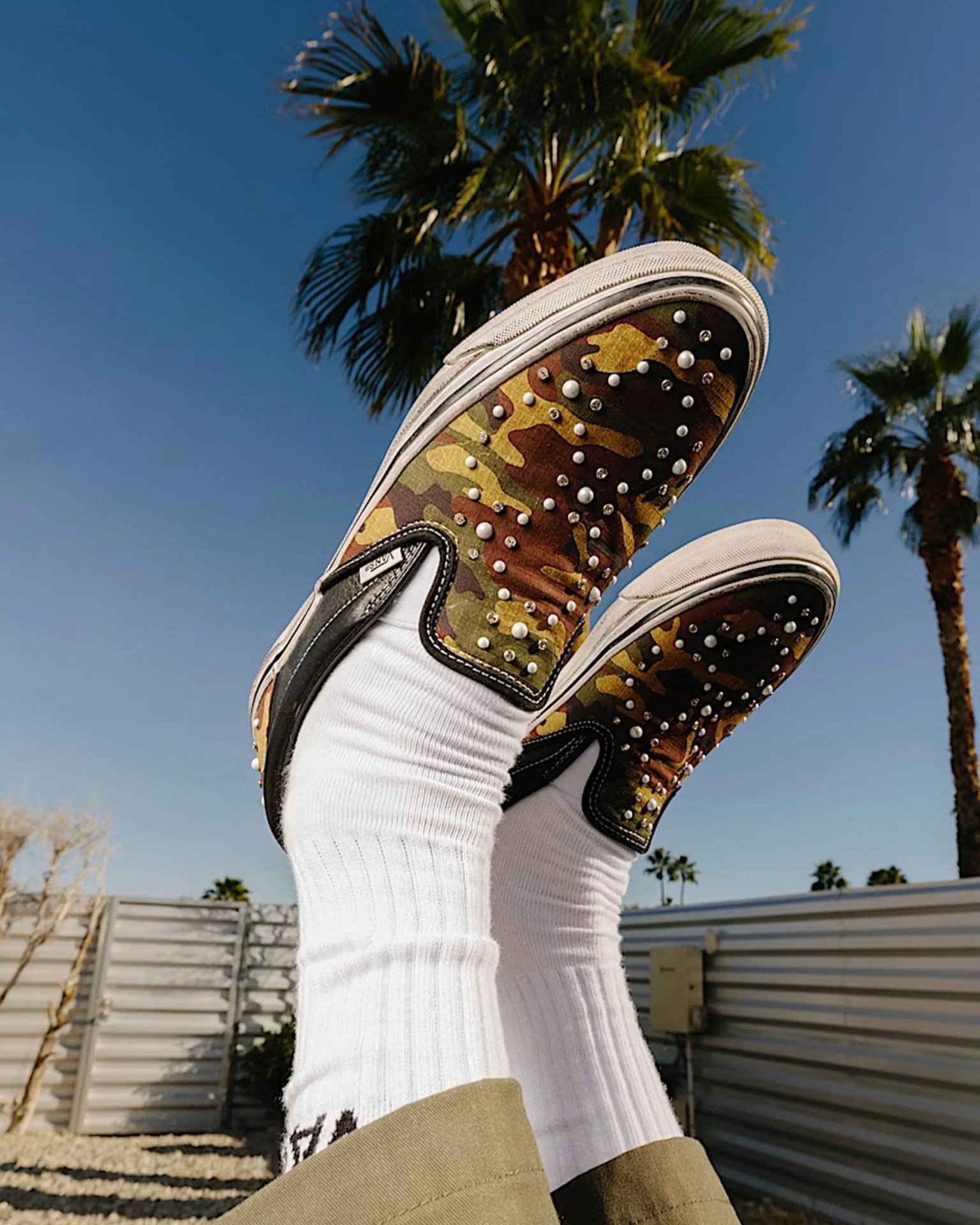 Pair of camo slip-on sneakers with pearl-like studs worn with white ribbed socks and khaki pants against a blue sky with palm trees.