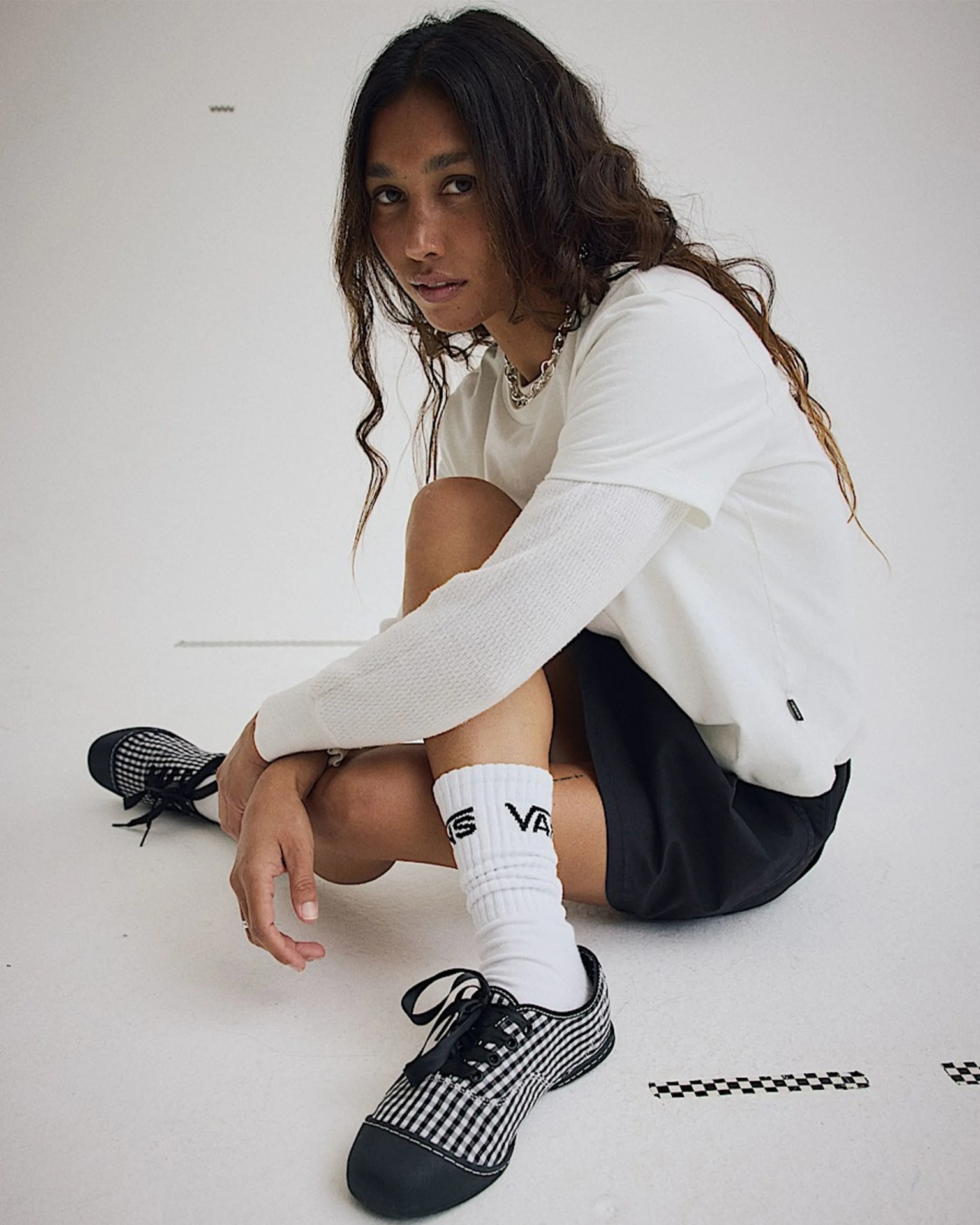 Person sitting on the floor wearing a white long-sleeve shirt under a white short-sleeve shirt, black skirt, white Vans socks, and black-and-white checkered Vans shoes.